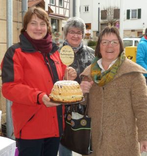 Gemeinderätin Roswita Decker-Röckel überreicht Bürgermeisterin Silke Höflinger zum 20-jährigen Marktstandjubiläum einen selbst gebackenen Gugelhupf, dahinter Mitbürgerin Frau Schliemann.